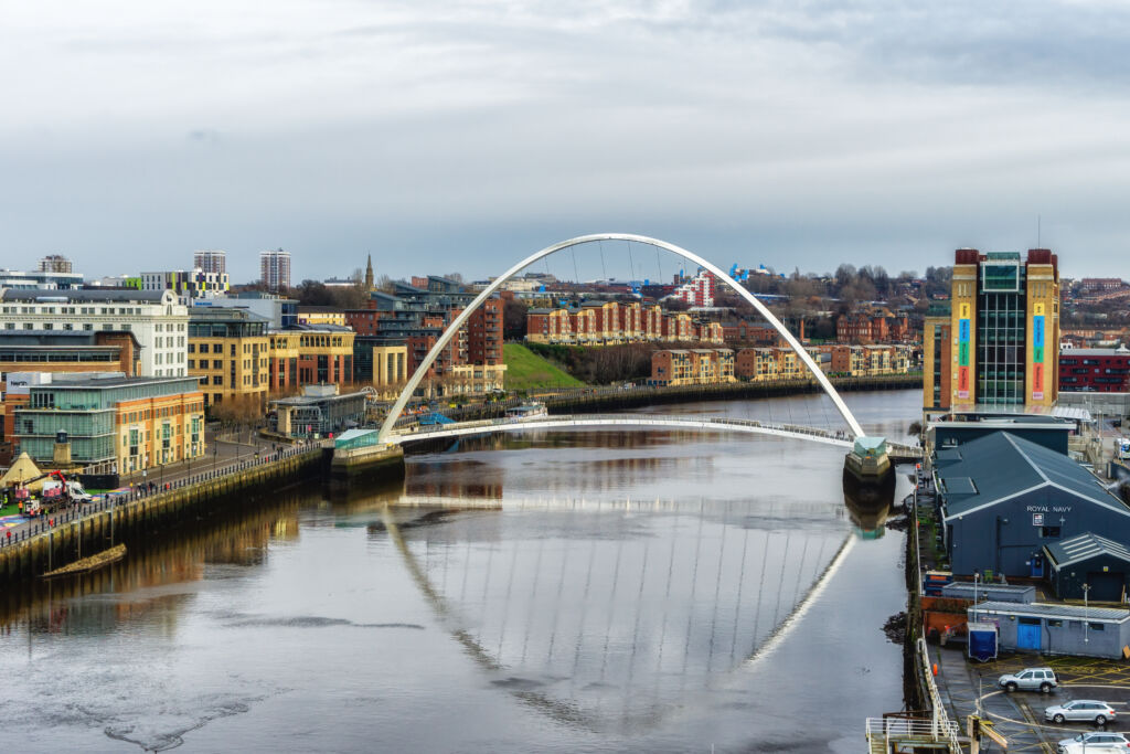 View of the River Tyne in Newcastle upon Tyne, showing the Gateshead Millennium Bridge in the foreground and the Tyne Bridge in the background. Buildings line both sides of the river, with quayside walkways, moored boats, and reflections on the water under a bright sky.