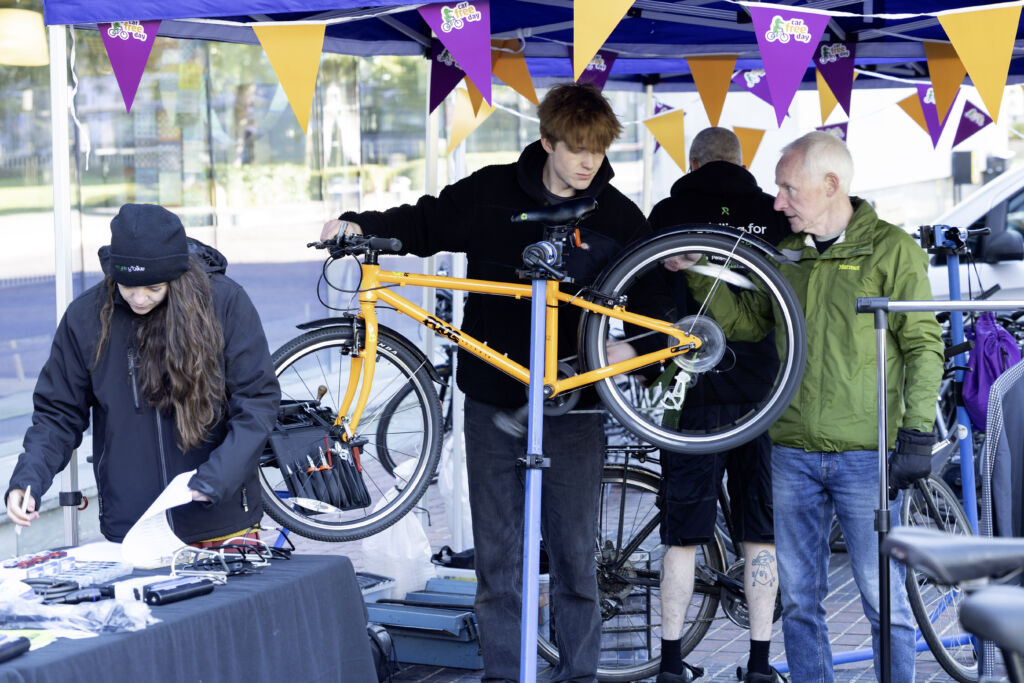 Several people are gathered under a blue canopy decorated with orange and purple “Car Free Day” bunting at a bicycle maintenance event. In the centre, a bright orange bicycle is mounted on a repair stand while two people work on the rear wheel. Another person stands at a table with paperwork and bike repair tools. Additional bicycles and repair equipment are visible in the background.